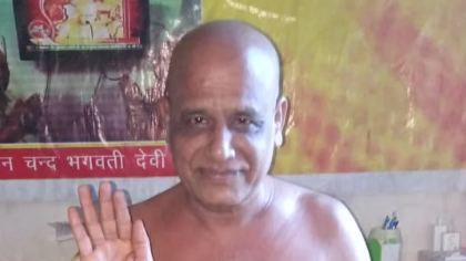 Bald man with bare shoulders smiling and waving his right hand in a colorful temple-like room.