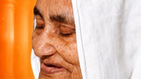 Close-up of an elderly woman with a white headscarf and freckles, eyes closed, against an orange background.