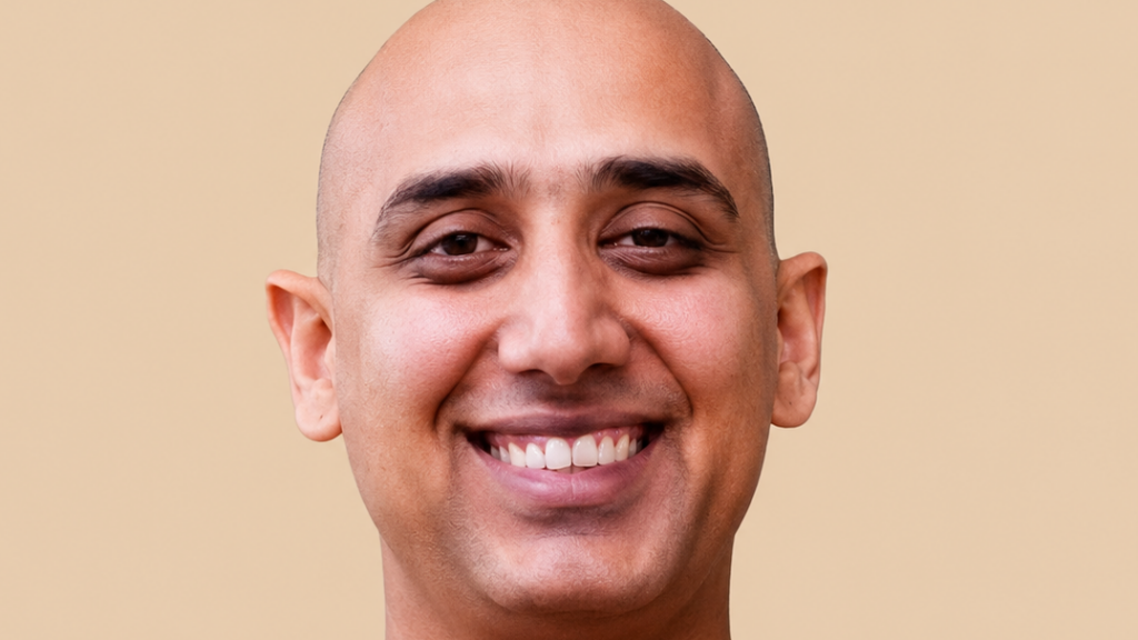 Close-up portrait of a smiling man with a shaved head on a beige background.