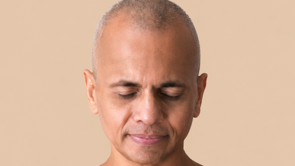 Close-up of a man with short gray hair and eyes closed, displaying a calm expression against a beige background.