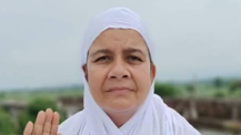 Woman in a white headscarf outdoors, raising her right hand in a greeting or blessing gesture.
