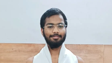 Portrait of a young man with a beard and glasses, wearing a white towel over his shoulders in a plain room.