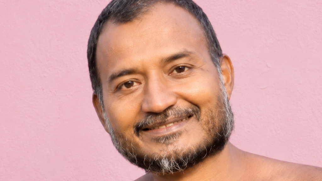 Smiling man with short dark hair and a gray beard against a pink wall.