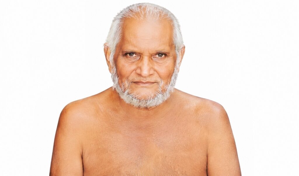Portrait of an elderly man with white hair and beard, facing the camera against a white studio background, shoulders bare.