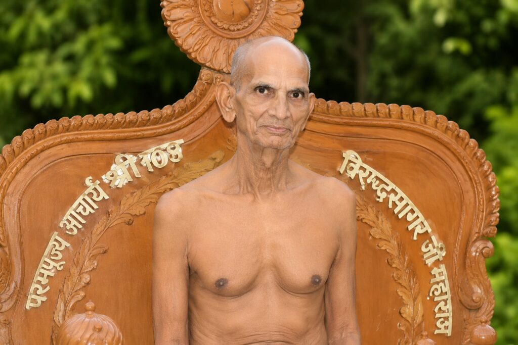 Elderly man, shirtless, sits on an ornate carved wooden throne with Sanskrit text, greenery behind.