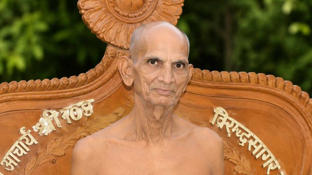 Elderly man seated shirtless on an ornate carved wooden throne outdoors, with Sanskrit inscriptions on the backrest.