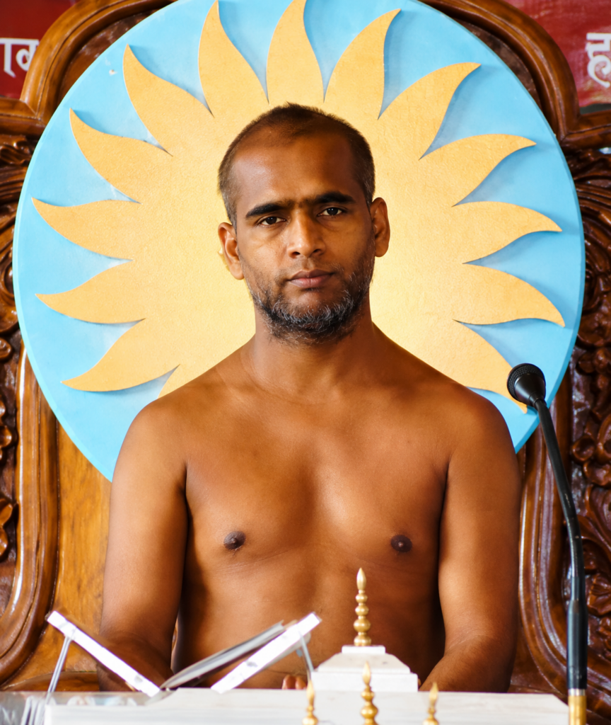 Shirtless man seated at a podium in front of a large blue circle with a yellow sunburst, microphone on his right side.