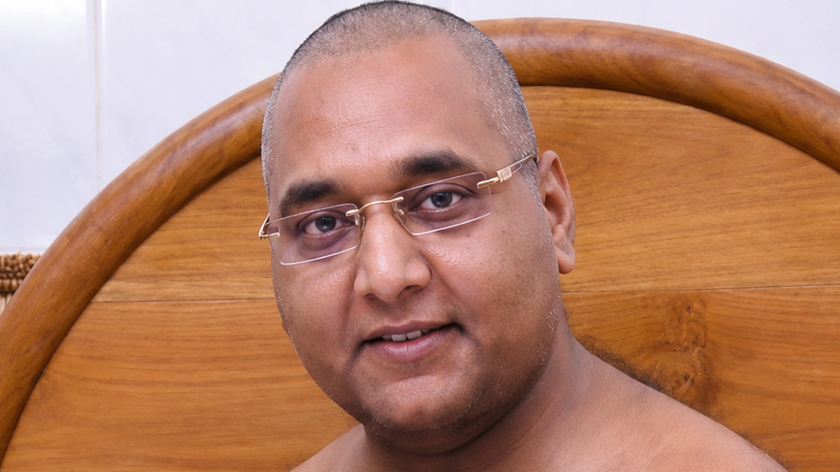 Close-up of a bald man wearing rimless glasses, facing the camera with a wooden headboard in the background.