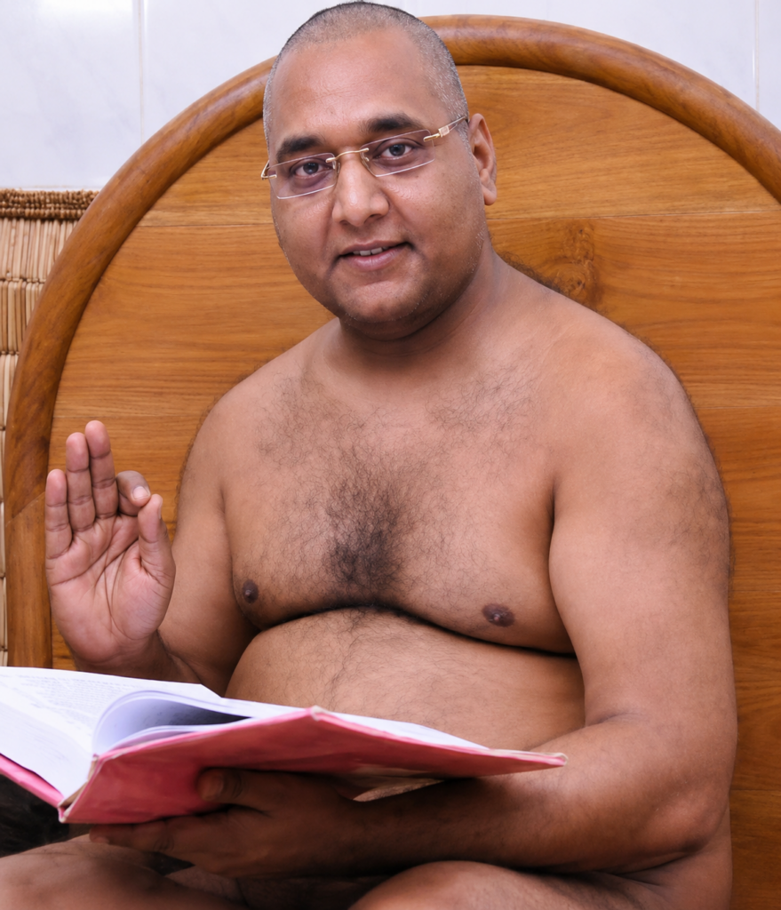 Shirtless man with glasses sits in bed against a wooden headboard, reading a pink-covered notebook and raising his right hand in a greeting gesture.