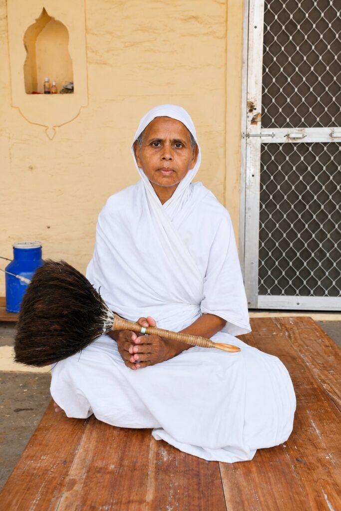 Woman in white robes sits cross-legged on a wooden bench, holding a broom in front of a beige wall and metal gate.