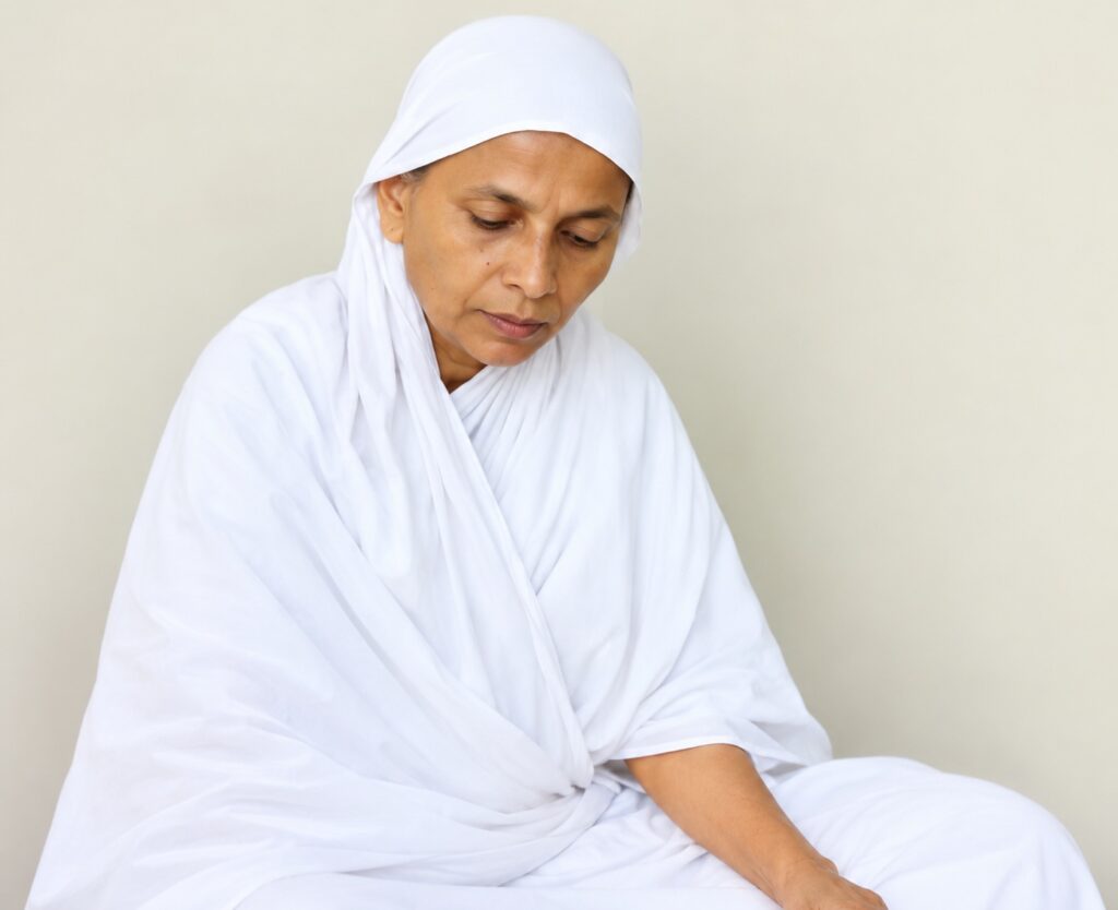 Woman in a white hijab and robe sits calmly, looking downward in contemplation.