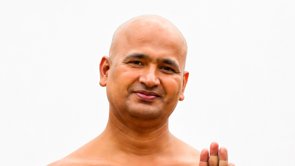 Smiling bald man with bare shoulders, raising one hand in a gentle greeting gesture.