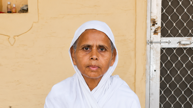 Elderly woman wearing a white headscarf and robe, standing against a yellow wall beside a metal mesh door.