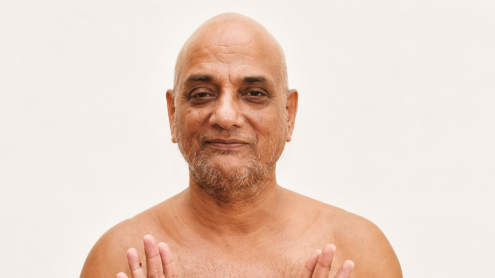 Smiling, bald man with a gray beard, shirtless, holding his hands up in a calm, greeting gesture against a light backdrop.