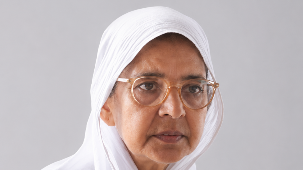 Portrait of an older woman wearing a white hijab and round glasses, looking slightly to the left against a gray background.
