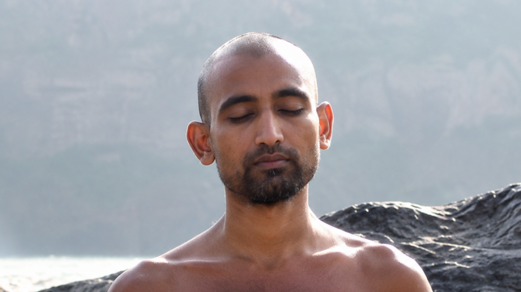 Man with a shaved head and beard meditating with eyes closed on a rocky shoreline by calm water.