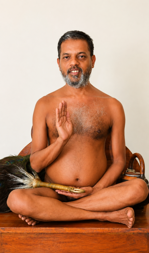 Bearded man sits cross-legged on a wooden surface, right hand raised in blessing while holding a feathered ceremonial whisk.