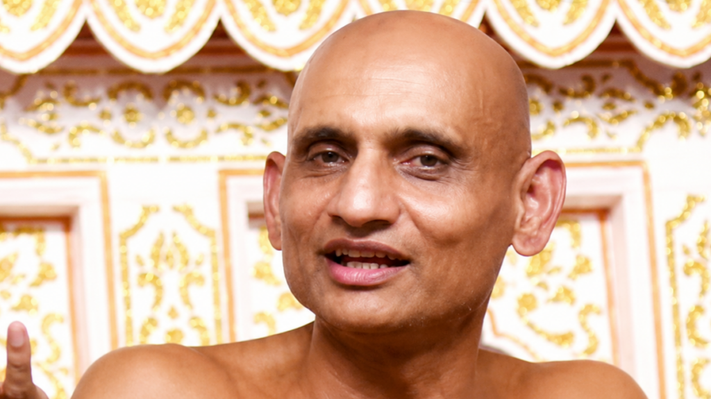 Close-up of a bald man speaking, ornate gold-patterned backdrop behind him.