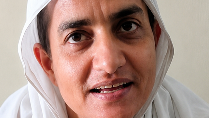 Close-up of a person wearing a white headscarf speaking to the camera, with a neutral background.