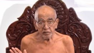 Older man with glasses sits shirtless in a large, ornate carved chair, gesturing with one hand toward the camera.
