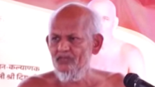 Close-up of an elderly man with a white beard speaking into a microphone at a public event.