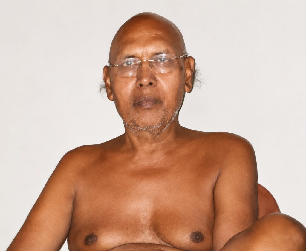 Older man with glasses, bald and lightly stubbled, shirtless against a pale background, looking at the camera.