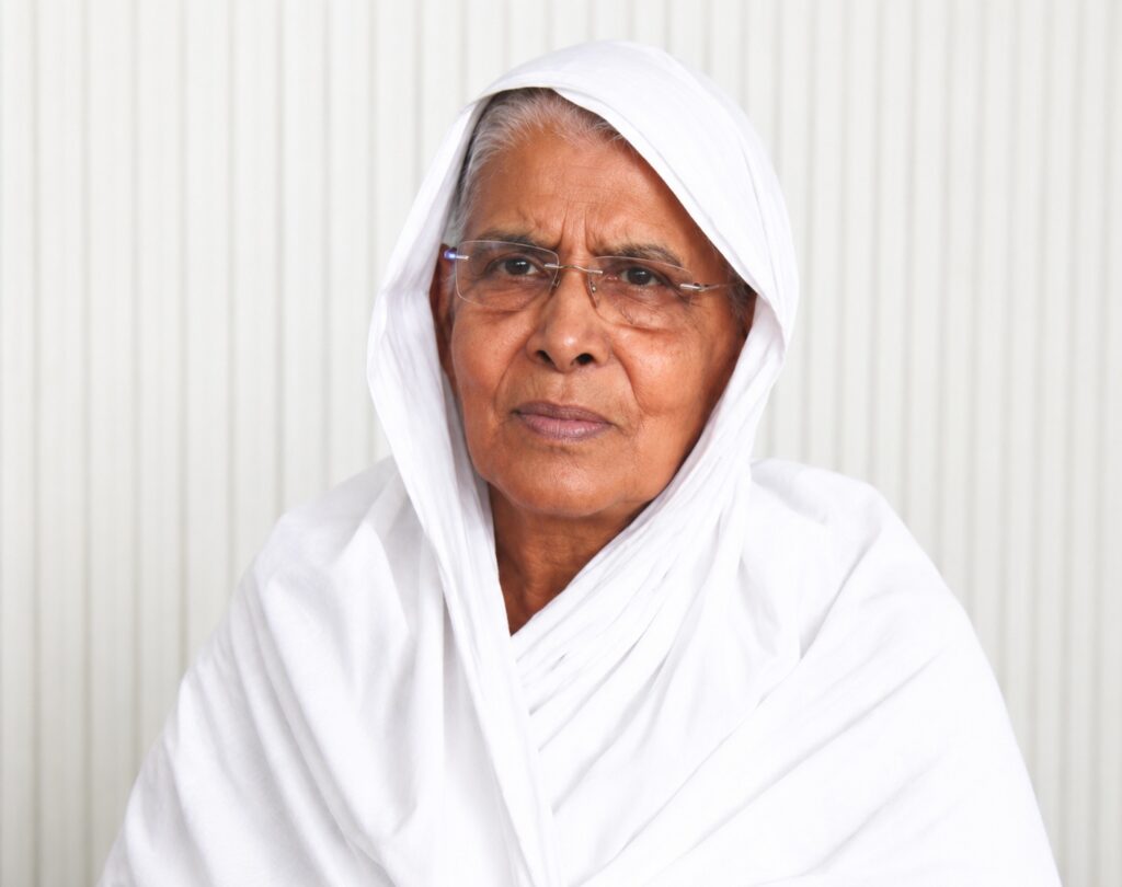 Portrait of an elderly woman wearing a white headscarf and glasses, looking at the camera.