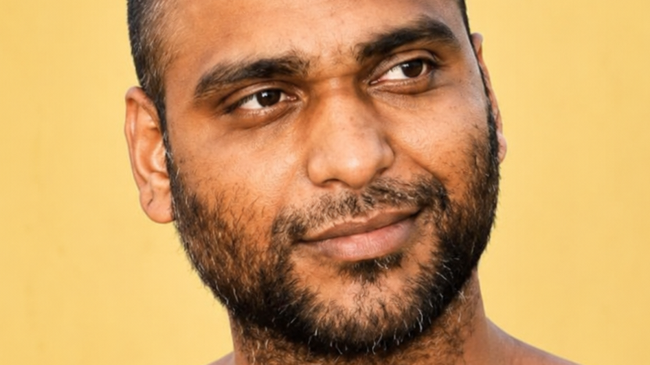 Close-up portrait of a man with short hair and a beard looking to the left against a warm yellow background.