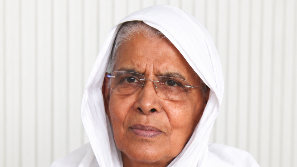 Close-up portrait of an elderly woman wearing a white headscarf and glasses, looking at the camera.