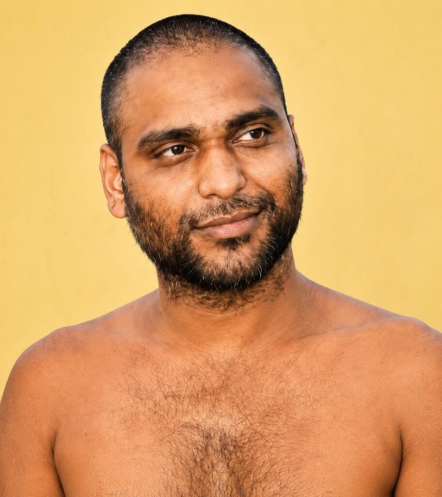 Portrait of a shirtless man with a short beard and mustache, looking to the right against a yellow background.