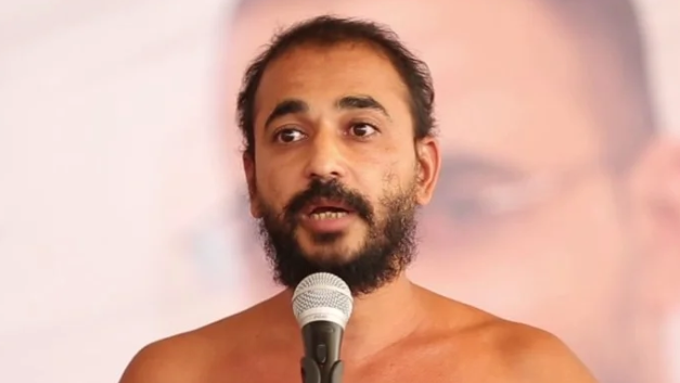 Bearded, shirtless man speaking into a microphone on stage.