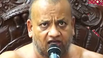 Close-up of a bearded, bare-chested man speaking into a microphone against an ornate carved wooden backdrop.