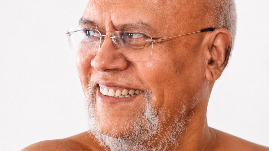 Smiling older man with glasses, gray beard, looking to the left against a light background.