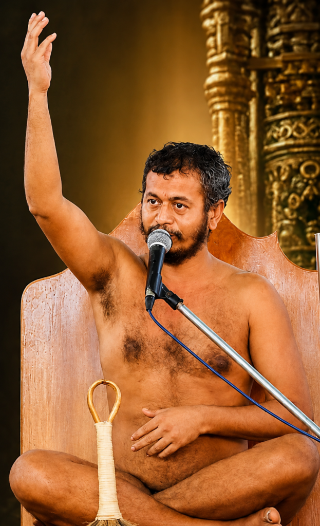 Shirtless man sits cross-legged on a wooden chair, speaking into a microphone with his right hand raised.
