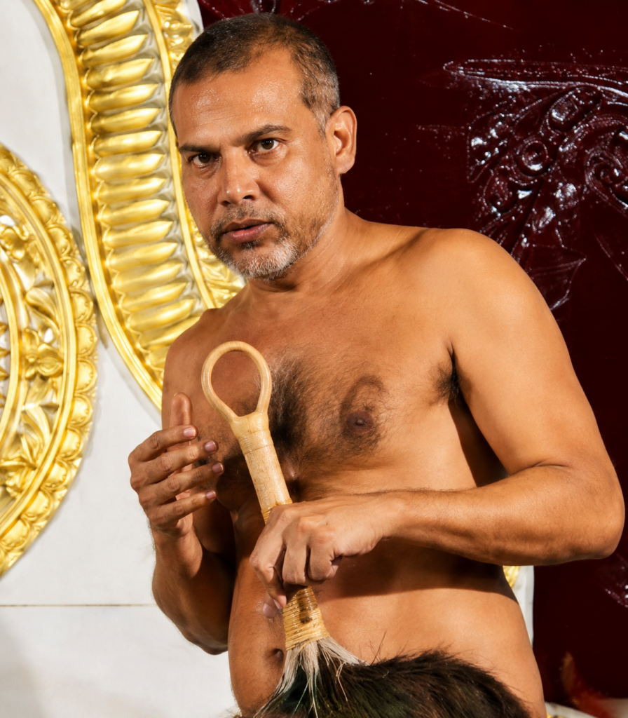 Middle-aged man, shirtless, holding a wooden ceremonial staff with a looped top, against an ornate gold frame backdrop.