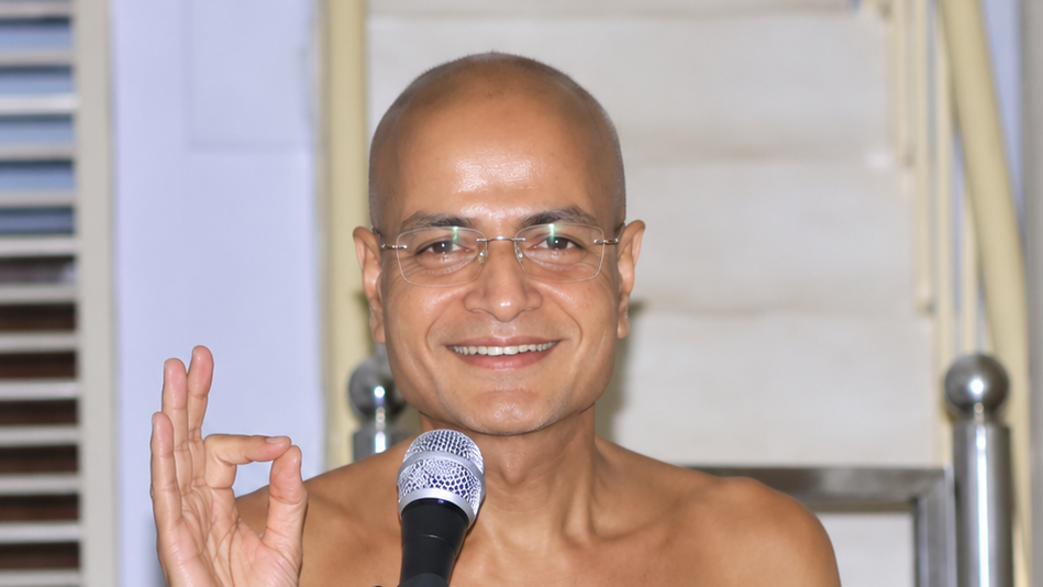 Bald man with glasses on stage, speaking into a microphone and making an OK sign with his right hand, smiling at the audience.
