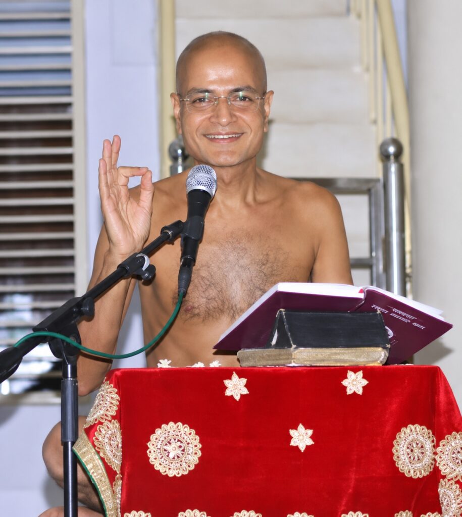 Smiling, bald man wearing glasses speaks into a microphone at a podium with a red patterned cloth and an open book in front of him.