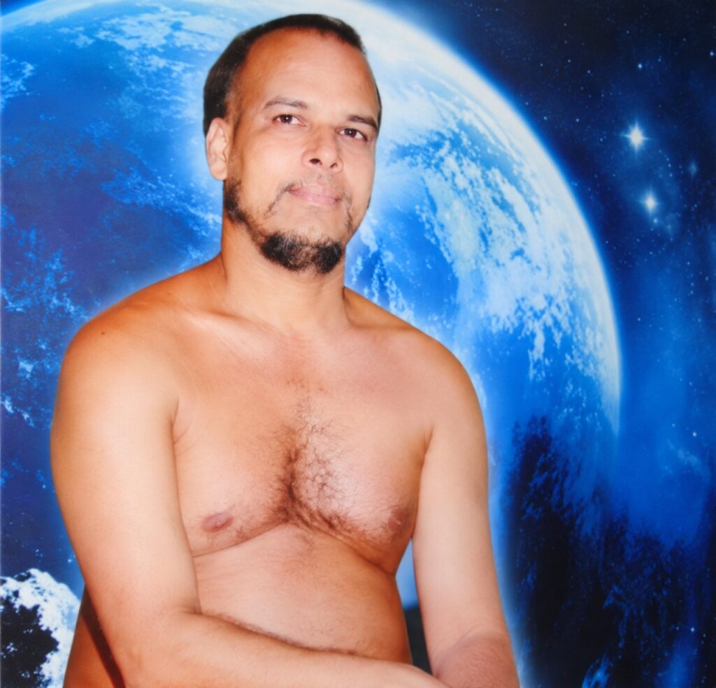 Man with a short beard poses shirtless against a space-themed backdrop featuring Earth and stars.