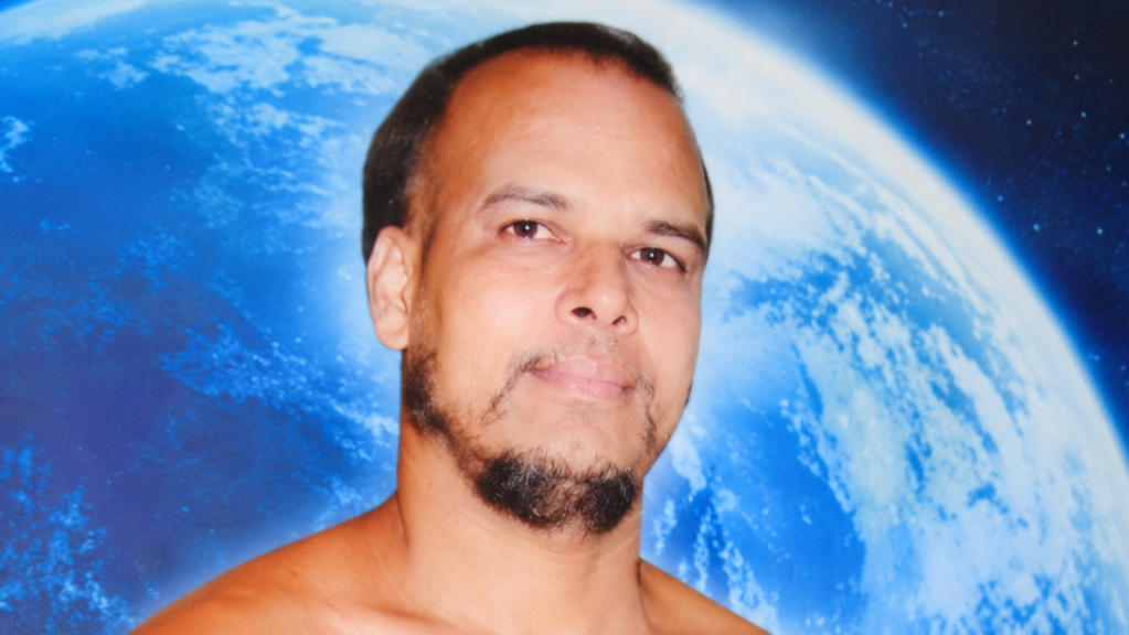 Man with short hair and goatee shirtless posing in front of a large Earth backdrop with blue space imagery