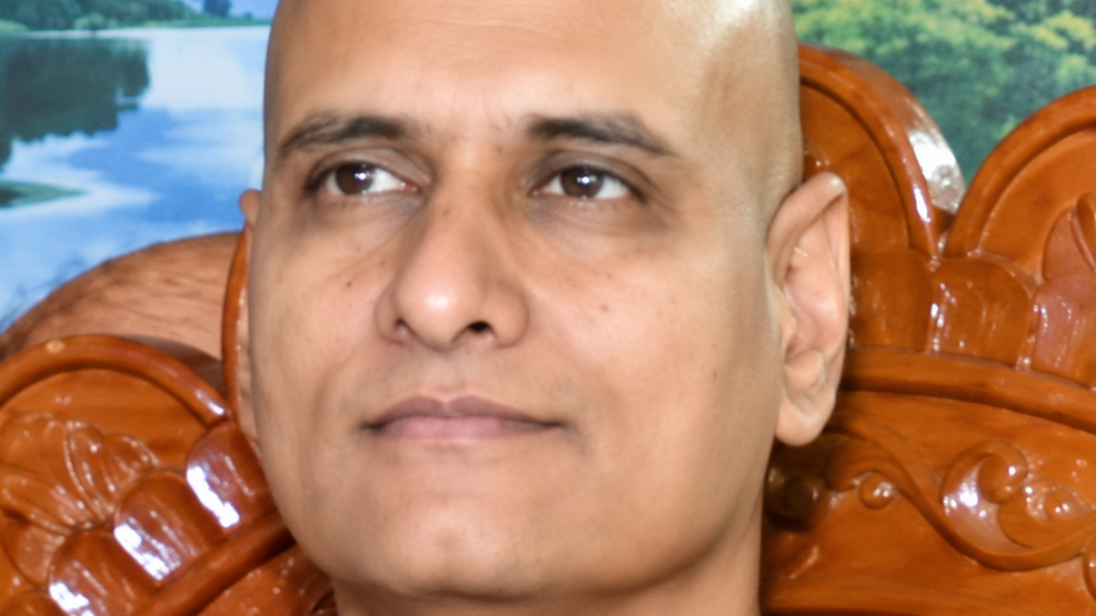 Close-up portrait of a bald man with a neutral expression seated on an ornate carved wooden chair behind him.