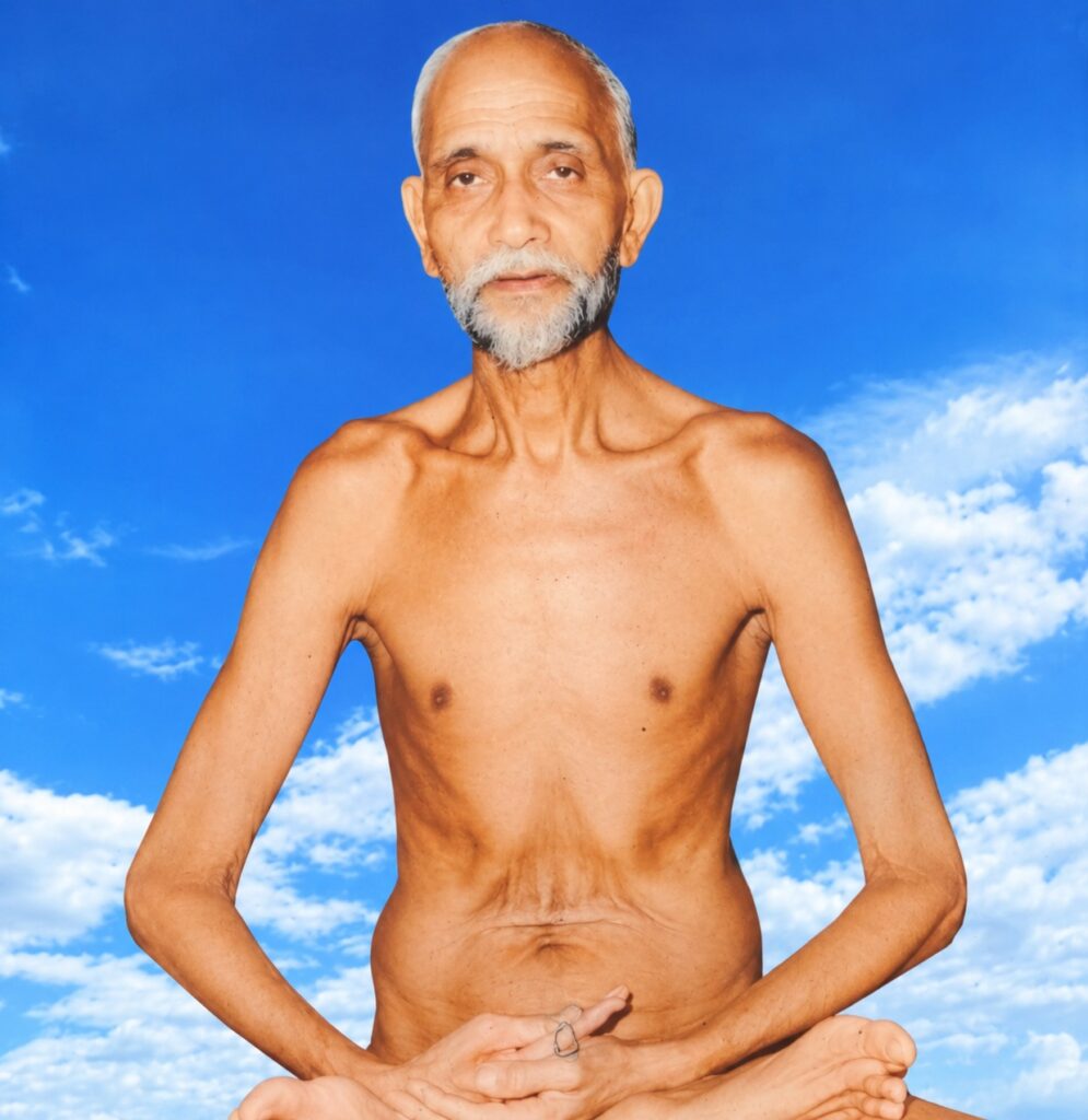 Older man with a gray beard meditating in a lotus pose against a bright blue sky with clouds.