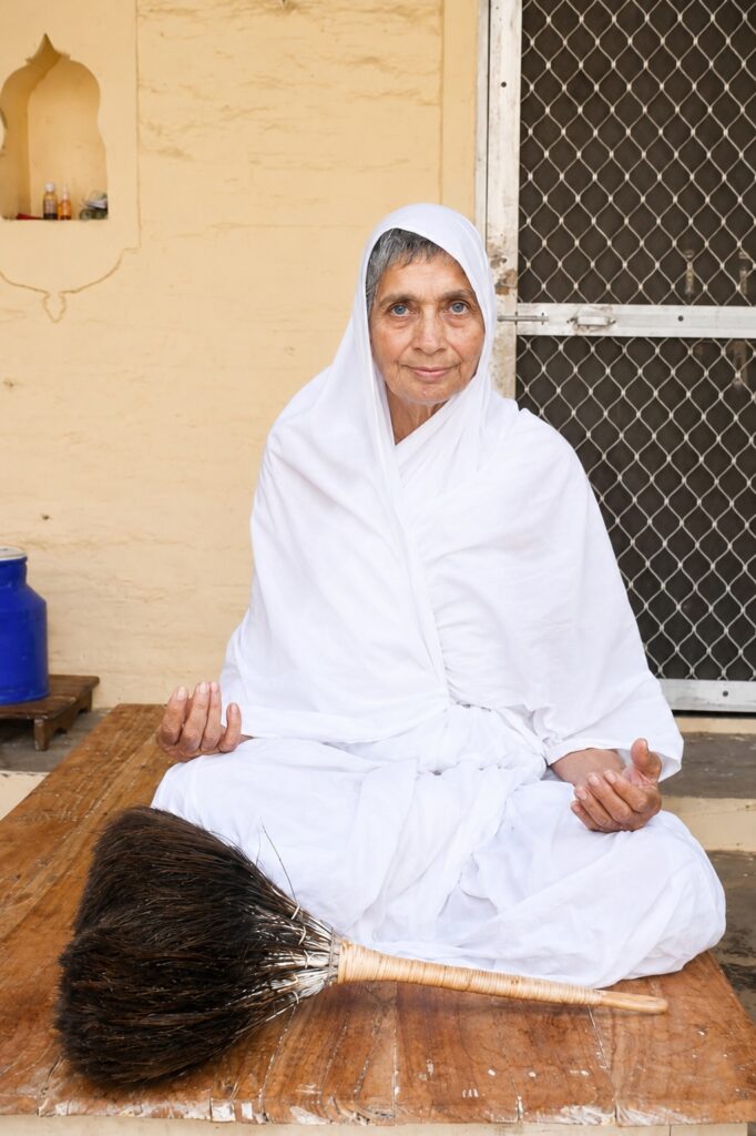 Elderly woman in a white robes sits cross-legged on a wooden bench outdoors, with a broom in front and a gated wall behind.