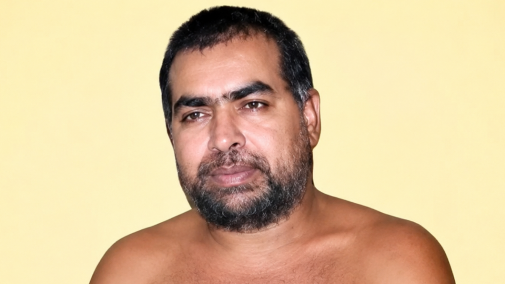 Portrait of a shirtless man with short dark hair and a full beard against a pale yellow background.