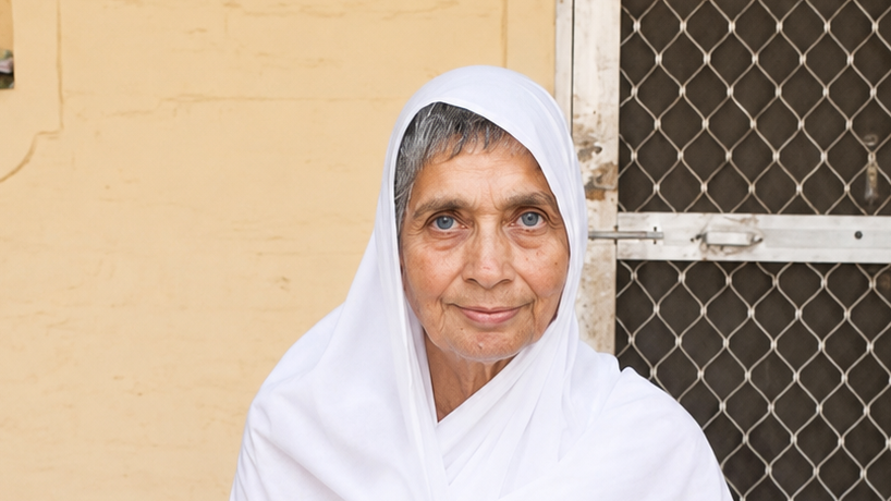 Portrait of an elderly woman in a white headscarf smiling at the camera, standing by a beige wall and a metal mesh door adjacent to her.