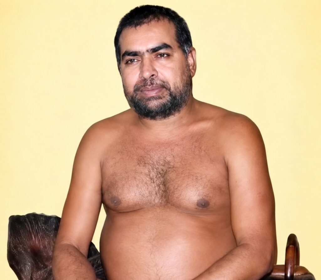 Man with short dark hair and beard, shirtless, seated against a pale yellow wall.
