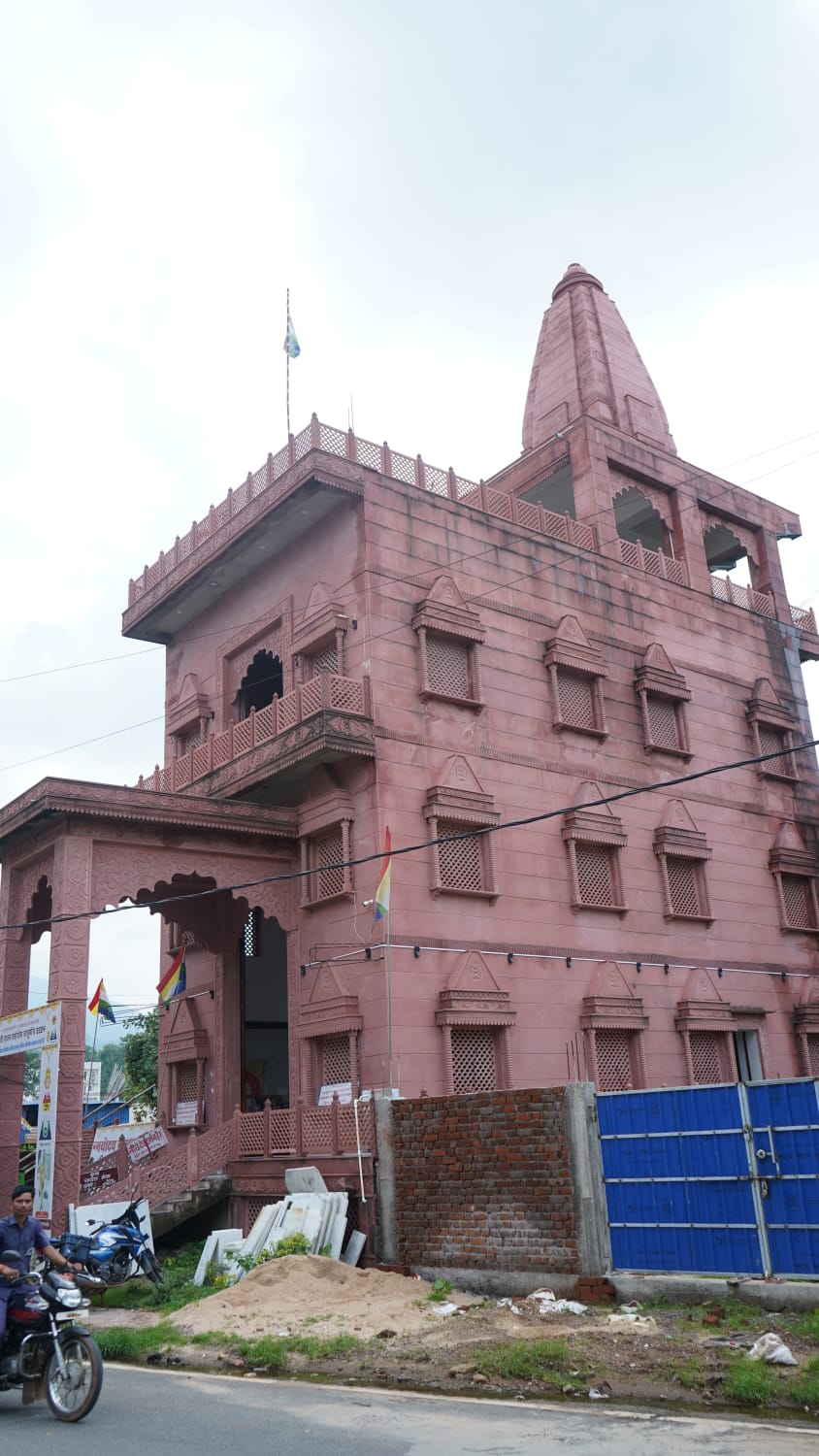 Pink temple-style building with intricate lattice windows and a small tower, flags at the entrance, under construction with bricks and a blue gate in the foreground.