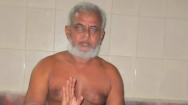 Older shirtless man with gray beard standing in a tiled room, hands pressed together in a greeting gesture (namaste).