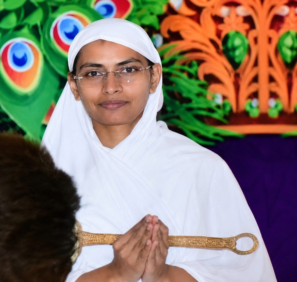 Young woman in white traditional attire with glasses, hands in prayer pose against a colorful stage backdrop.