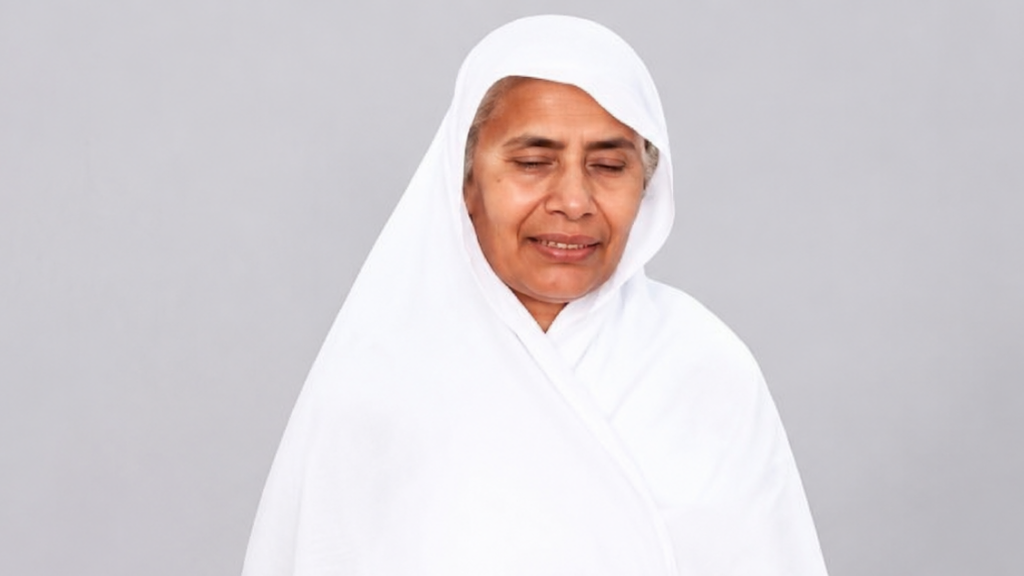 Older woman in a white headscarf and robe, eyes closed, smiling softly against a gray background.