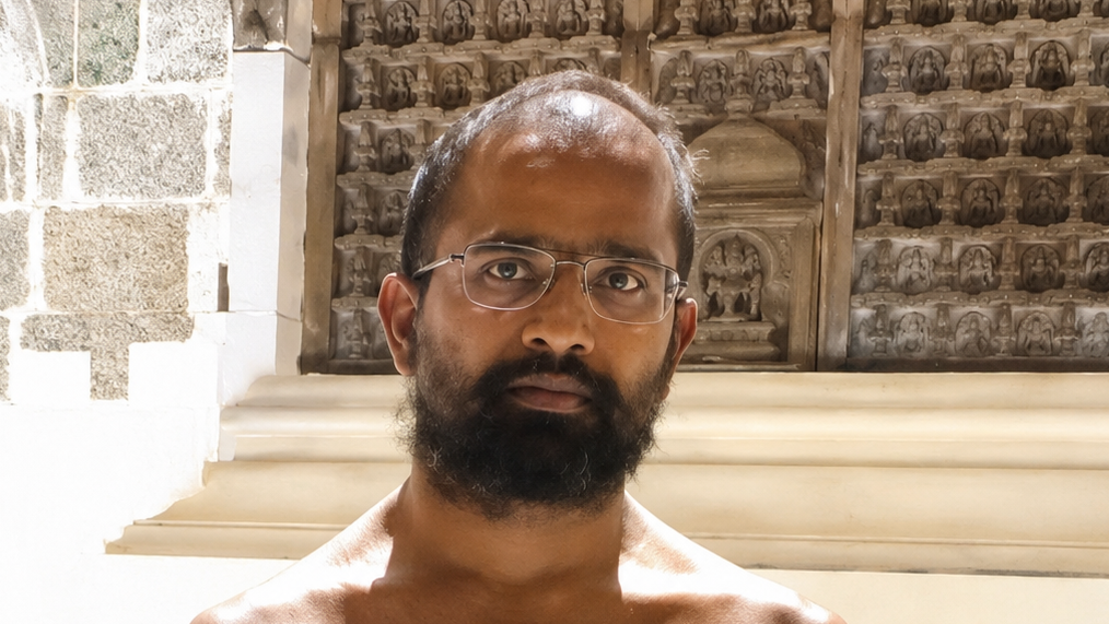 Man with glasses and a beard standing shirtless in front of a carved temple wall, looking at the camera.